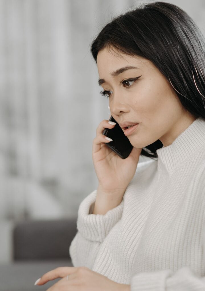 Focused woman talking on phone while typing on laptop indoors, showcasing remote work or online communication.