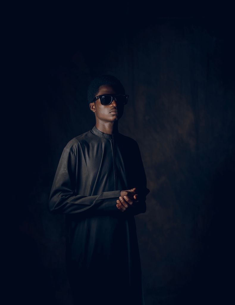 Portrait of a stylish man wearing dark sunglasses and a traditional outfit against a moody background.