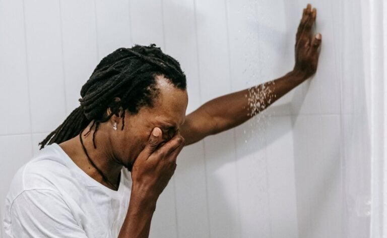 Side view of anonymous sorrowful young African American guy with dreadlocks covering face with hand and crying while standing in shower cabin