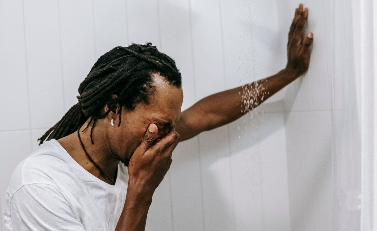 Side view of anonymous sorrowful young African American guy with dreadlocks covering face with hand and crying while standing in shower cabin