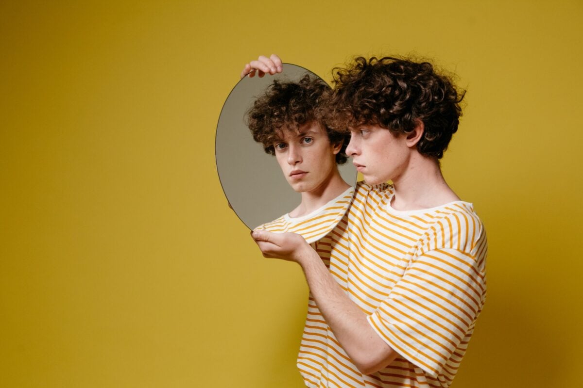 Studio portrait of a young man holding a mirror against a yellow background.