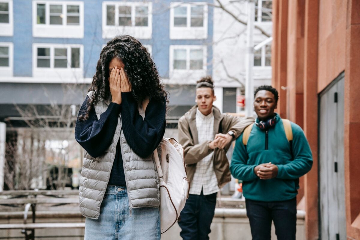 Unrecognizable young female student covering face with hands while being bullied by multiethnic male classmates on street