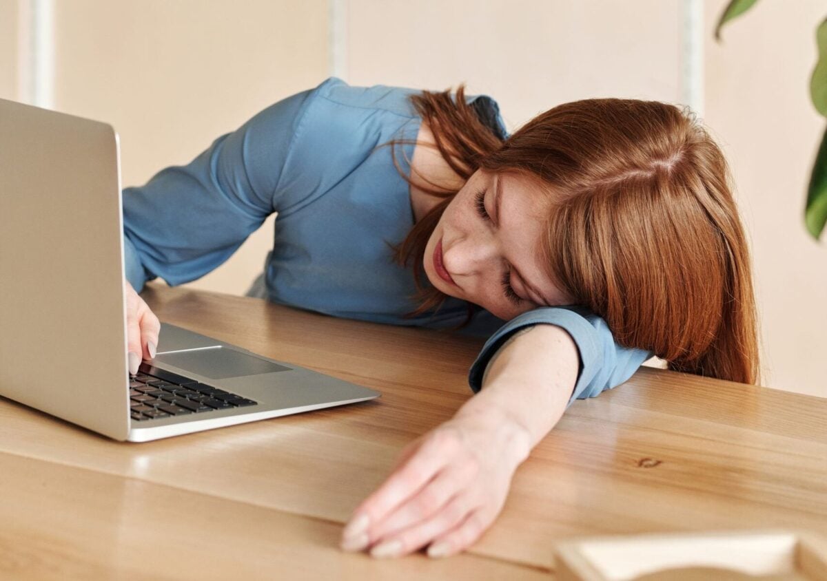 Woman in blue dress asleep on desk with laptop nearby in a modern office.