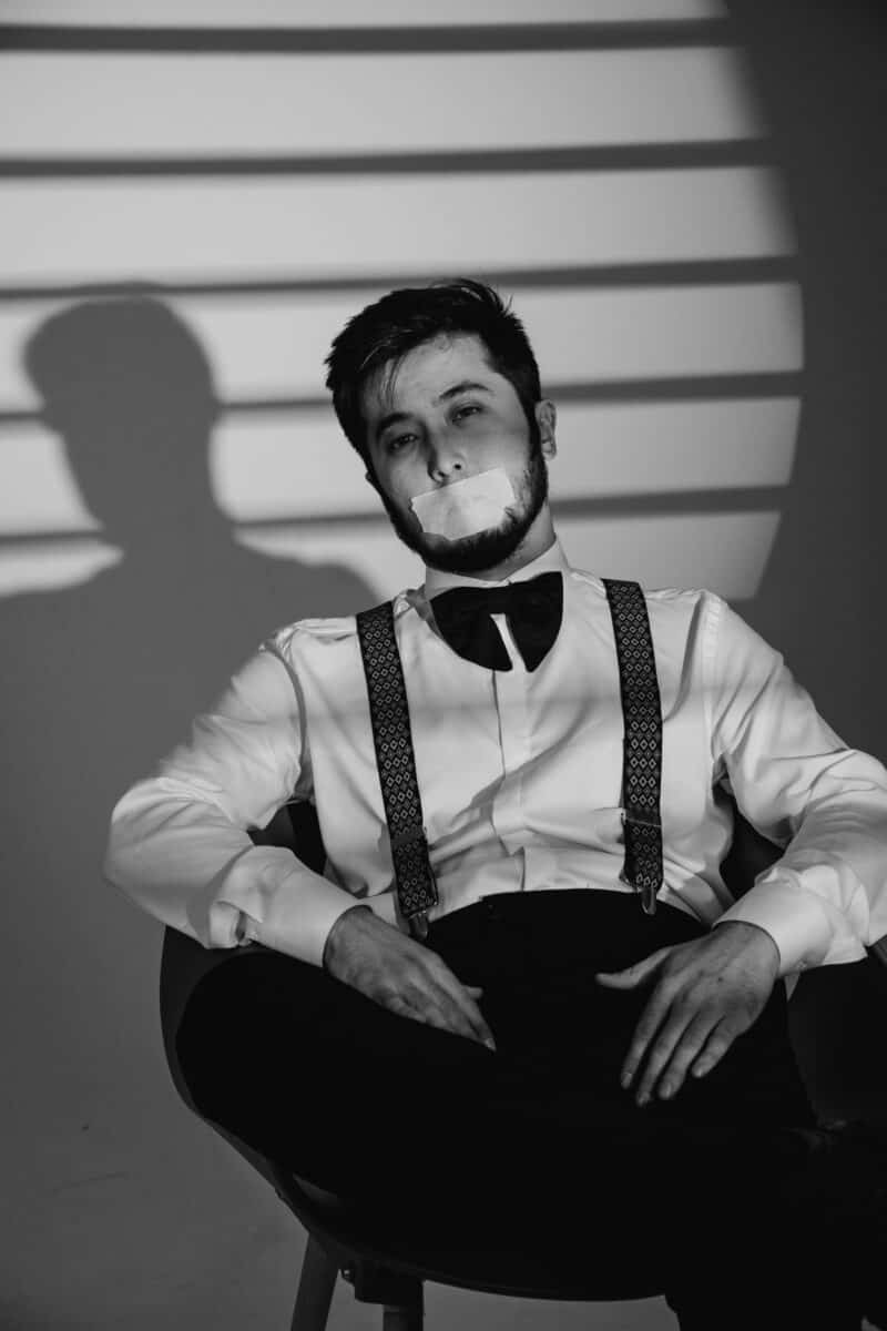 A black and white portrait of a man with taped mouth, sitting in a chair, wearing a bow tie.