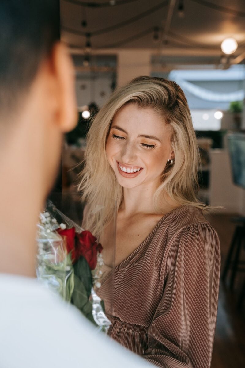 A joyful young woman with blonde hair receives red roses, smiling warmly indoors.