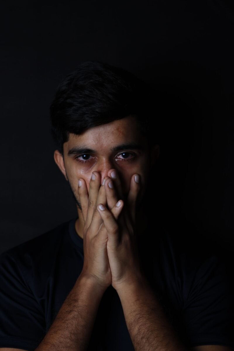 A man in a dark setting with hands over mouth, showing intense emotions.