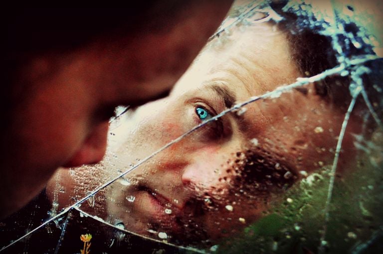 A man with green eyes peers into a cracked, wet mirror, conveying solitude and thought.