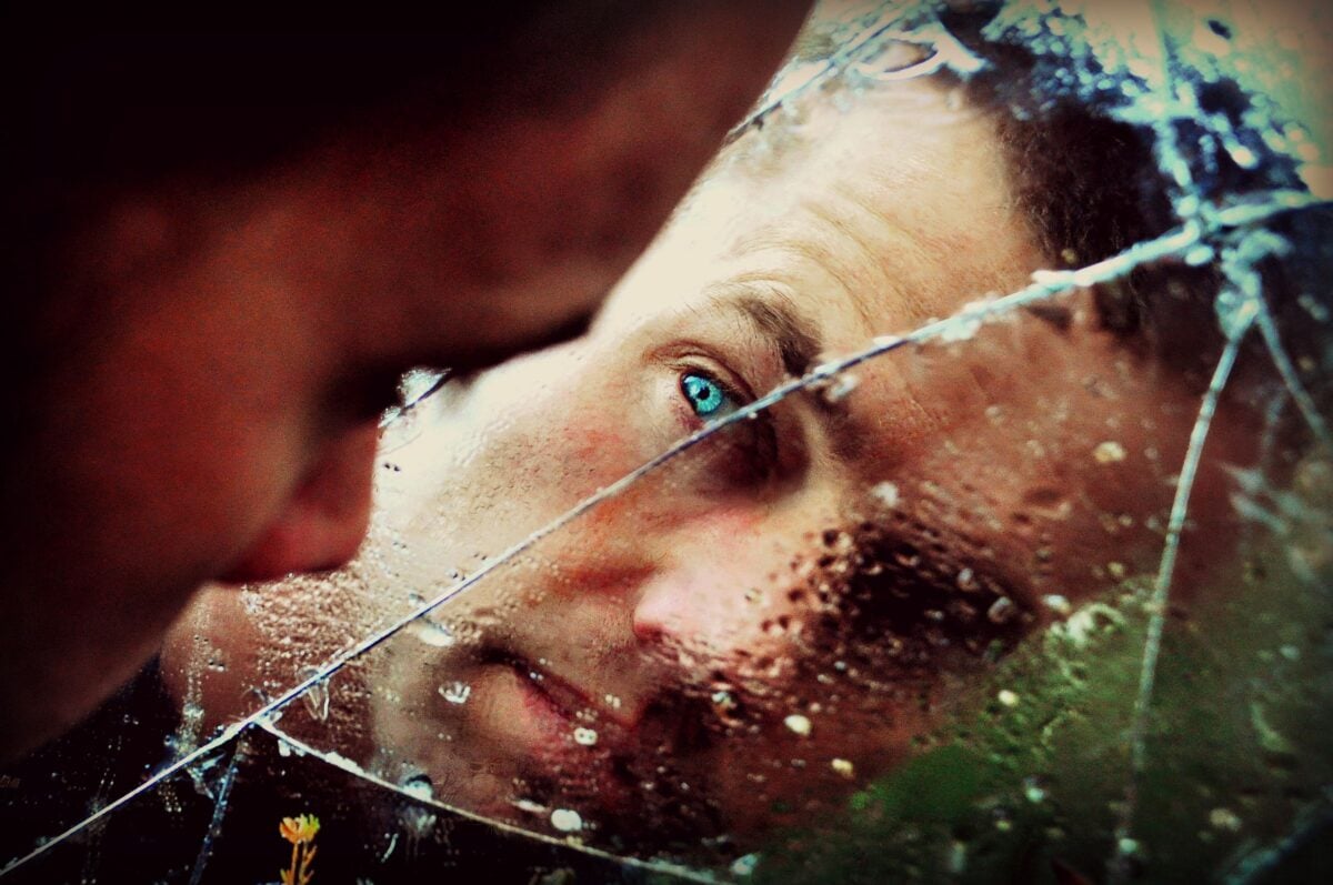 A man with green eyes peers into a cracked, wet mirror, conveying solitude and thought.