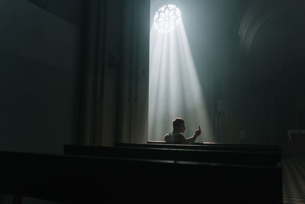 A person sits praying inside a church, illuminated by rays of sunlight through a window.