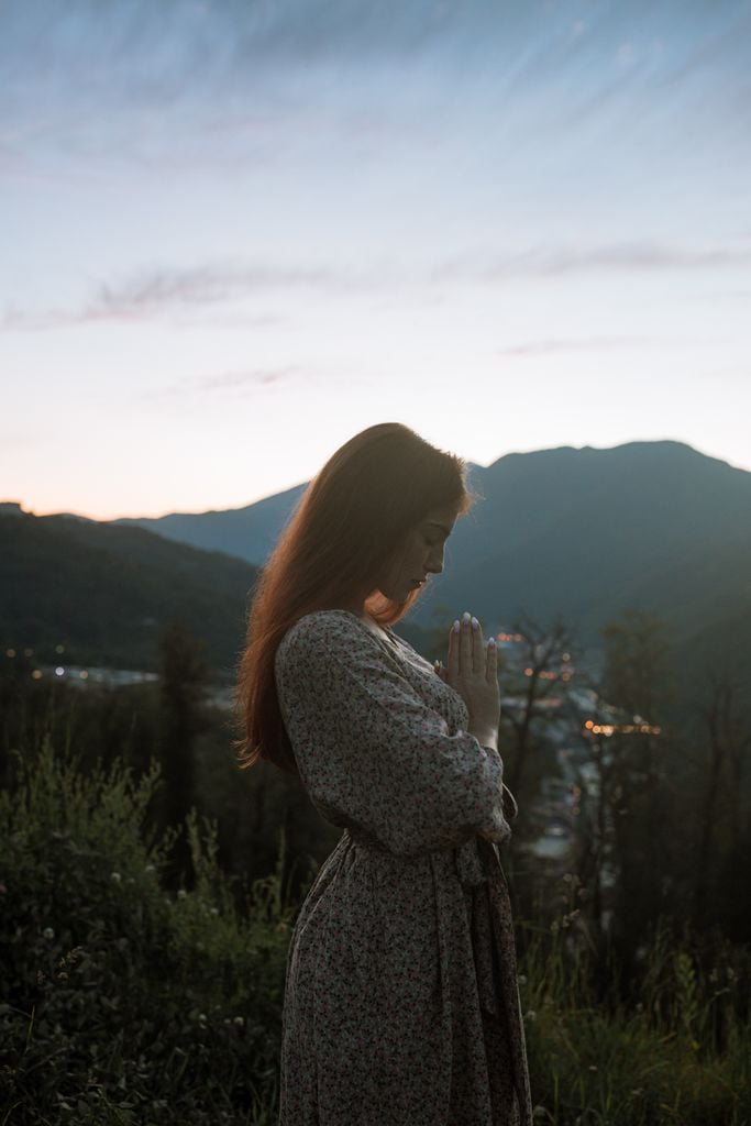 A serene portrait of a woman praying outdoors with mountains in the background.