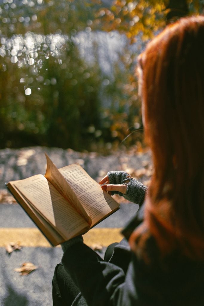A woman reads a book outdoors in an autumn park with sunlight filtering through the trees.