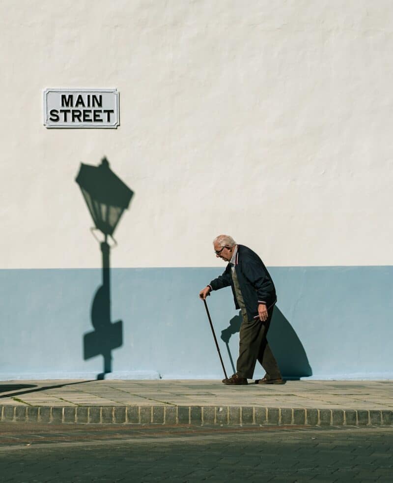 An elderly man with a cane walking along the Main Street sidewalk in Gibraltar, beside a classic street lamp shadow.