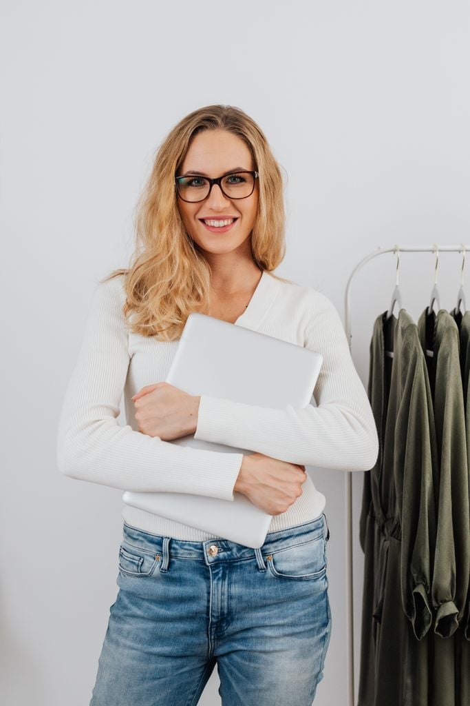 Blonde woman in casual wear with laptop, standing near clothing rack, smiling indoors.