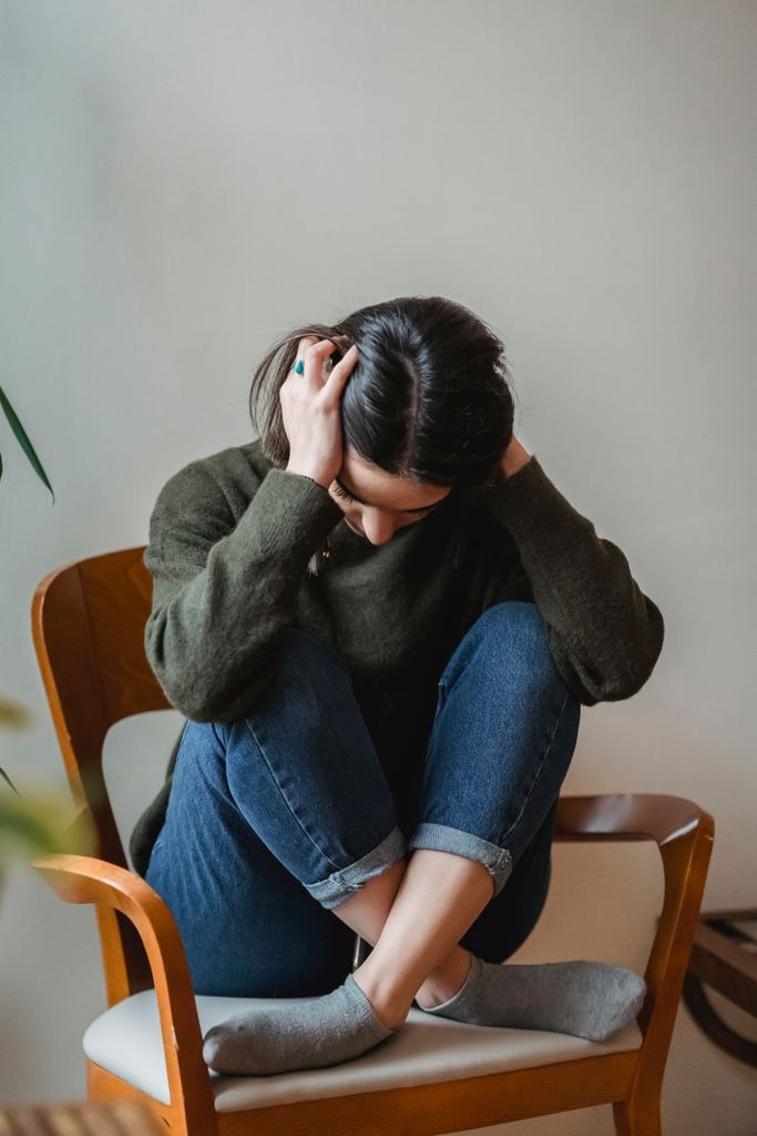 Full body of nervous depressed young female with dark hair in casual clothes covering ears with hands while sitting on chair with crossed legs
