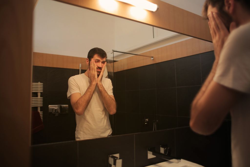 Low angle of exhausted male in casual outfit standing in front of mirror at home and touching weary face while suffering from headache