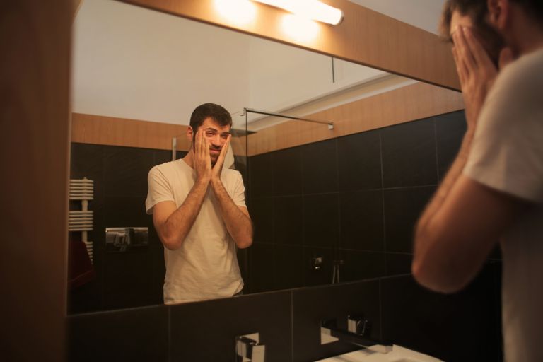 Low angle of exhausted male in casual outfit standing in front of mirror at home and touching weary face while suffering from headache