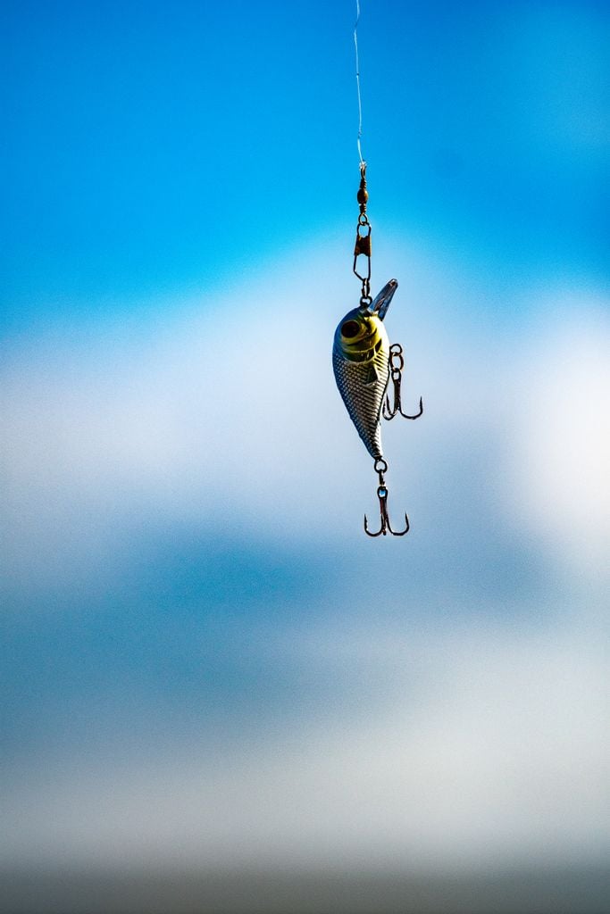 Metallic fishing lure with hooks suspended in mid-air against a bright blue sky.