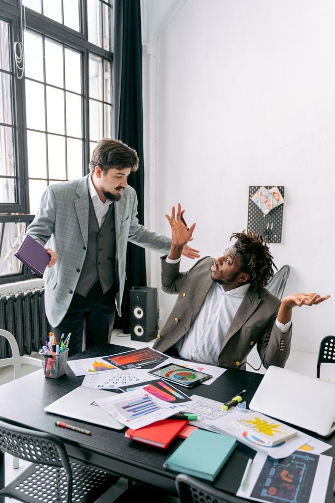 Two male colleagues in an office engaging in a tense discussion over papers and a tablet.