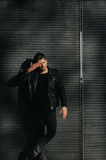 Young man poses confidently in a black leather jacket against a gridded metal urban wall.