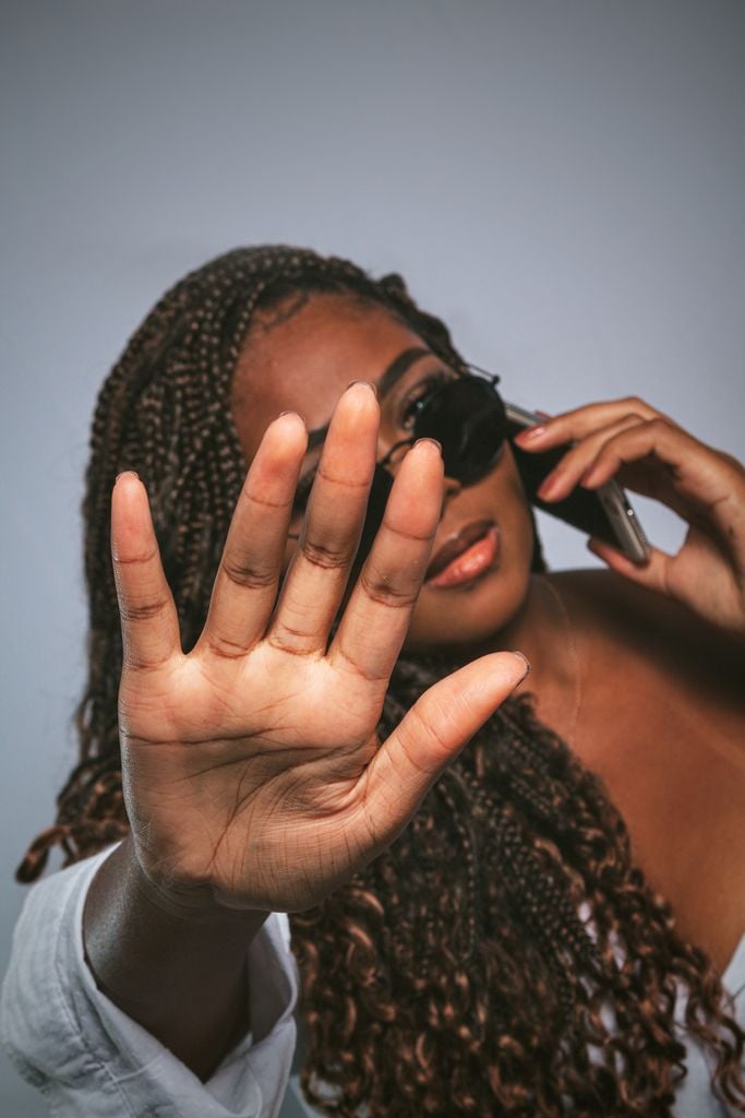 Young woman in sunglasses using a phone and gesturing to stop. Fashionable and assertive portrait.