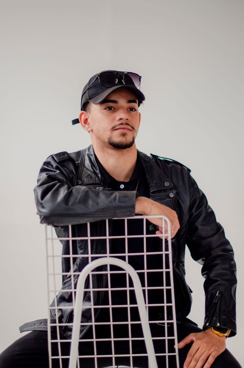 A confident man posing with a chair in a leather jacket and cap, exuding modern style.
