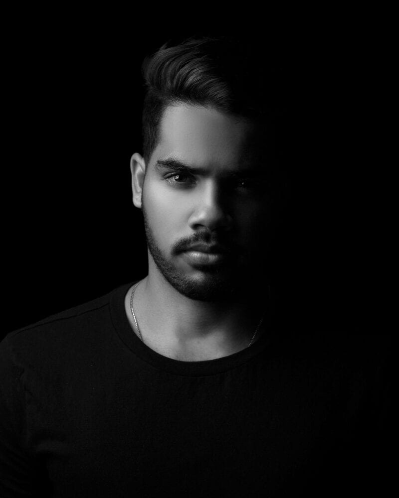 Black and white of serious young bearded ethnic male model in casual clothes looking at camera with concentration in dark studio
