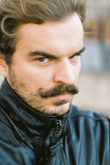 Close-up portrait of a man with a serious expression and mustache, wearing a jacket outdoors.