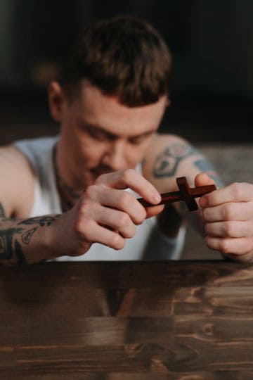 Tattooed man praying with a wooden crucifix inside a dimly lit church, symbolizing faith and reflection.