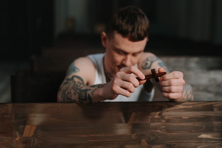 Tattooed man praying with a wooden crucifix inside a dimly lit church, symbolizing faith and reflection.