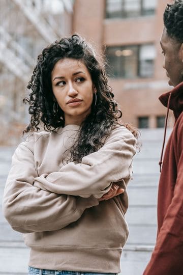 Unset ethnic woman with crossed arms and African American man looking at each other while having quarrel on street with building on blurred background